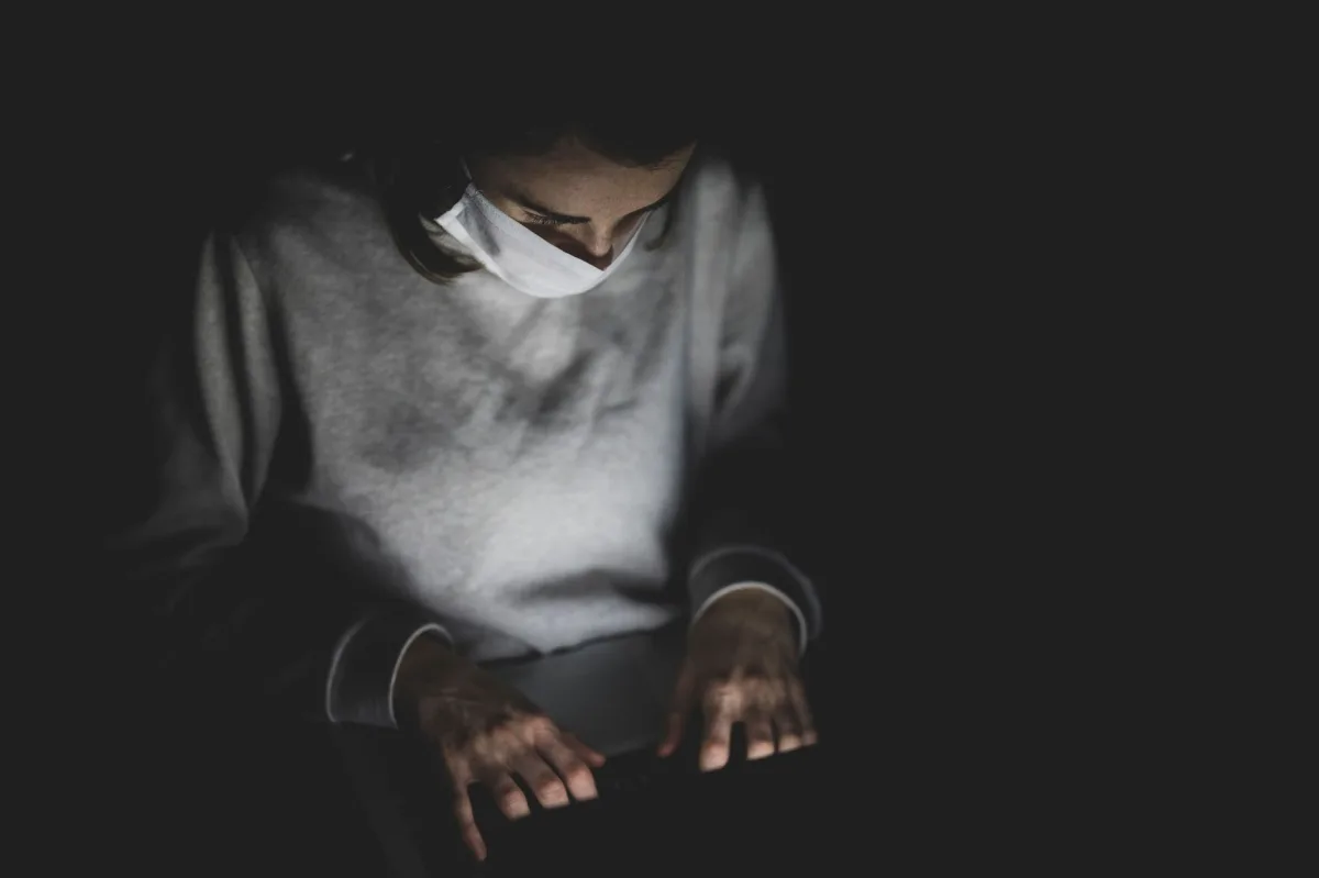 A person wearing a face mask works on a laptop indoors, showcasing pandemic isolation.