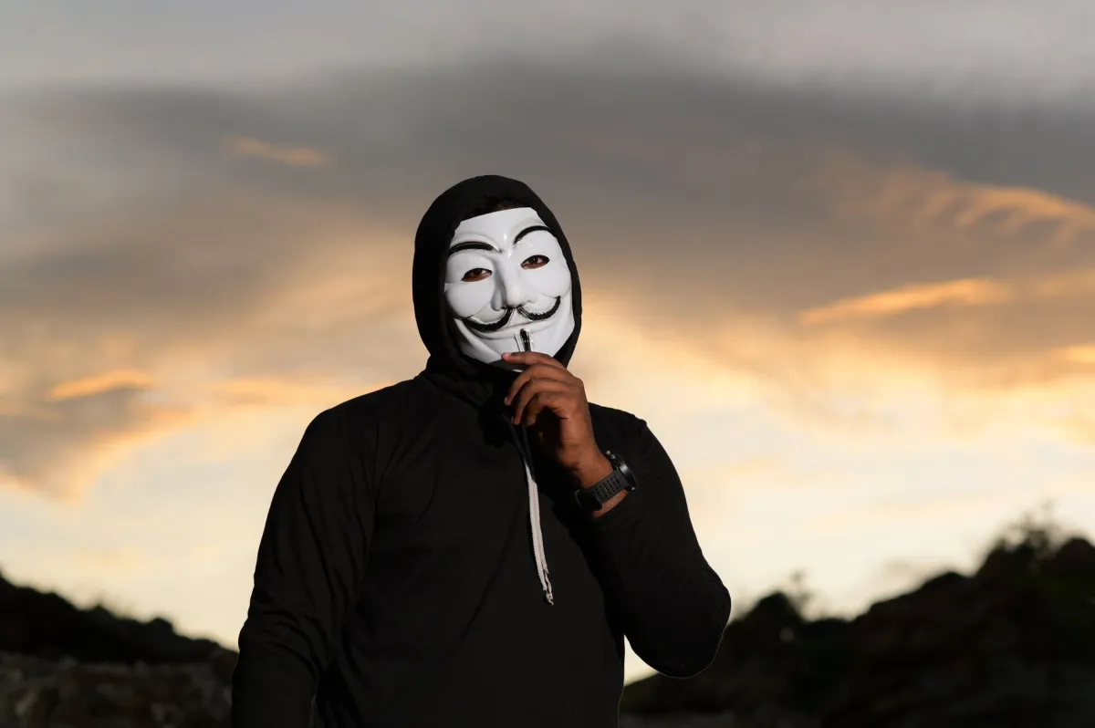 A person in a Guy Fawkes mask stands outdoors at sunset, exuding mystery.