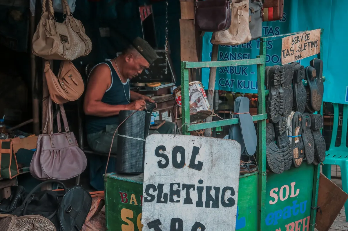 A local vendor repairing shoes at a bustling marketplace stall with visible signs and merchandise.